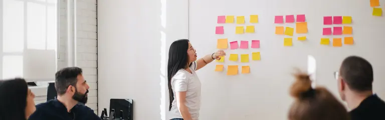 People having a meeting around a table, standing woman pointing to sticky notes on wall
