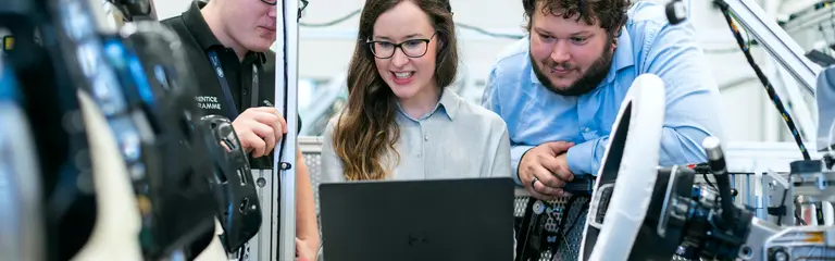 Group of three people, 2 men and 1 woman in the middle, looking at a laptop, surrounded by tech.