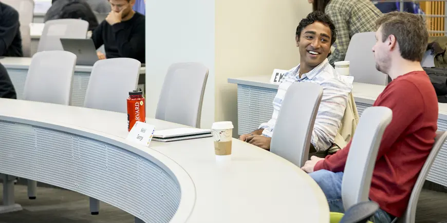 Nikhil sitting in class with a classmate. They are facing each other and talking. There are other students in the background taking their seats. 