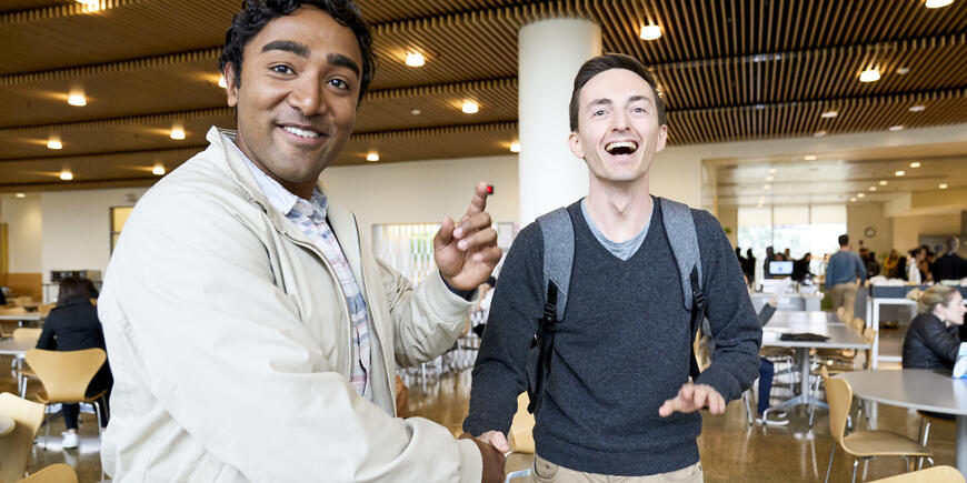 Nikhil and a friend smiling at the camera while shaking hands in the Sloan cafeteria