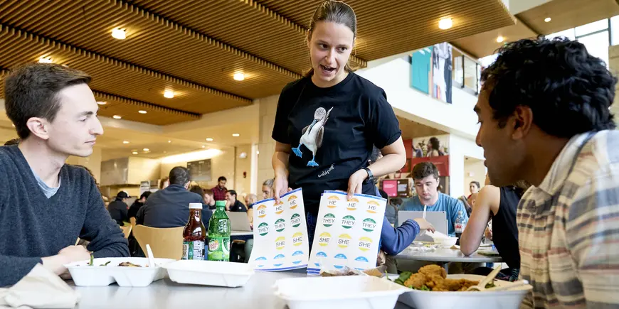 Emily standing over Nikhil and his friend having lunch. She is holding up stickers sheets that read "He", "She", "They". In the background, other students having lunch in the cafeteria.  