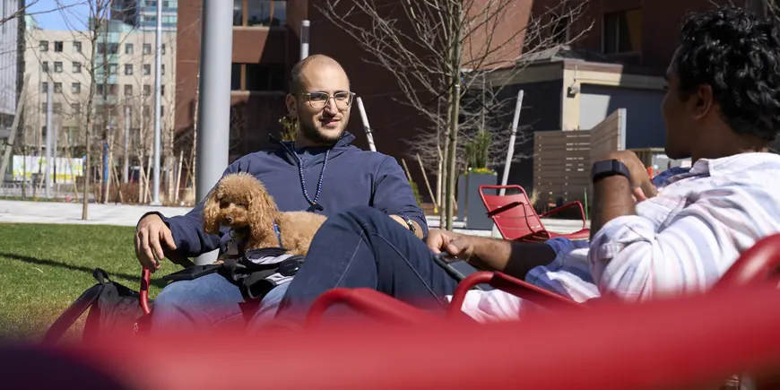 Nikhil and his friend sitting next to each on red lawn chairs. Sitting on his friend's lap is a small, cute poodle. 