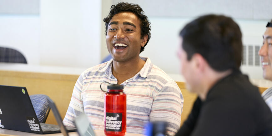 Nikhil smiling, in the foreground, his red water bottle and laptop screens