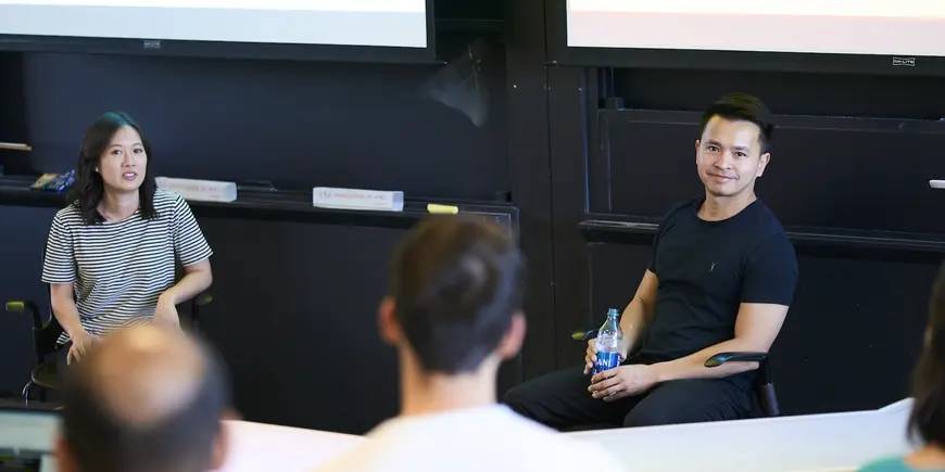 Two people sitting in office chairs facing a classroom filled with students. In the background two projector screens. 