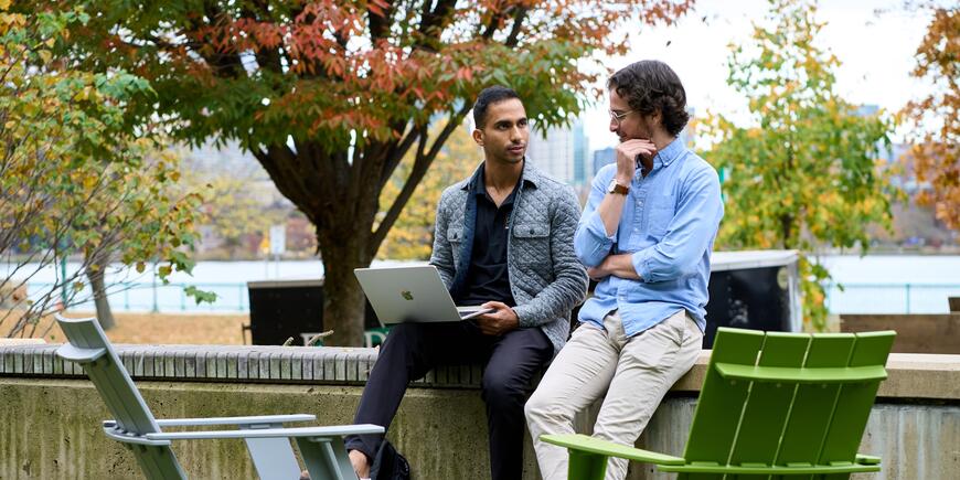 LGO student Abhi Rao sits outside with a laptop, in conversation with another student.