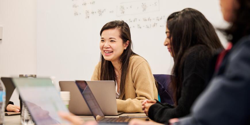 MBA student Sharon Fan sits at a table with other students. They are talking and looking at laptops.