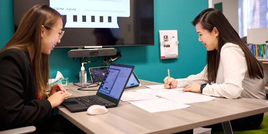 MBA students Sharon Fan and Gloria Wu work together over papers and a laptop.