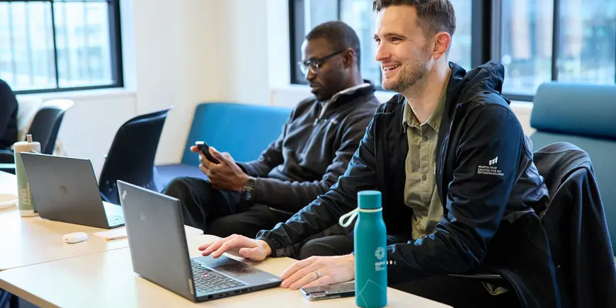 David Brown sits at a long table with another person. He looks ahead, smiling, with a laptop in front of him. 