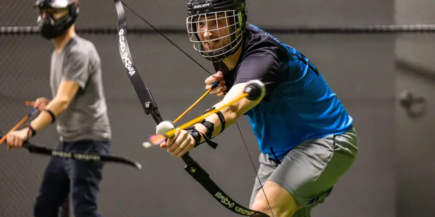 David Brown wears athletic wear and stands in an indoor athletic facility. He wears a helmet and holds a bow and an arrow with a soft, round ball on the end. He draws back the arrow as if to fire. 