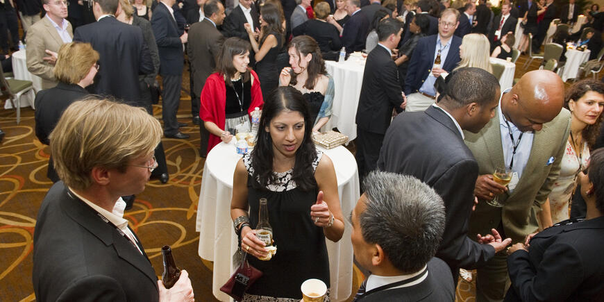 A woman speaks to two men in foreground of large social gathering