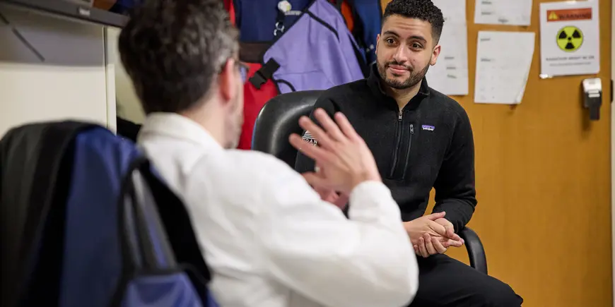 Mike Sanchez sits in an office in a hospital conversing with someone in a white lab coat. 