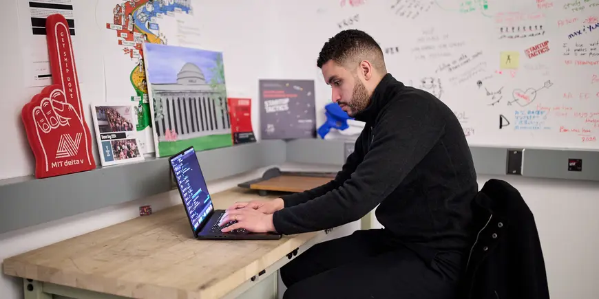 Mike Sanchez sits at a desk in a room in the Trust Center. He is looking at a laptop and typing. 