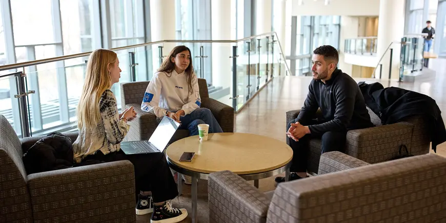 Mike Sanchez sits at low chairs in the MIT Sloan building with two other MBA students. 