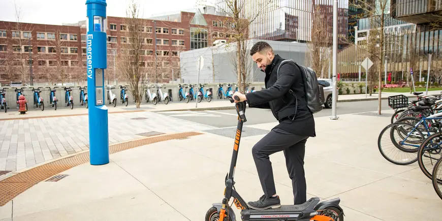 Mike Sanchez stands on a street corner with an electric scooter. 