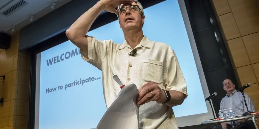 A man with his hand raised above his eyes looking out from stage
