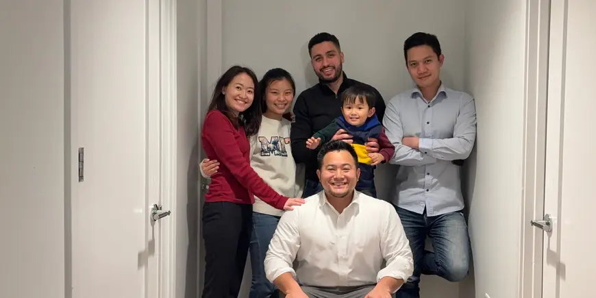Mike Sanchez poses for a photo. There is one person to his left, two to his right, and one on a chair in front of him. Mike Sanchez also holds a smiling toddler up to the camera. 