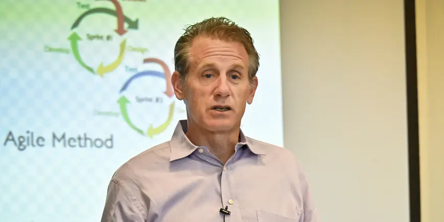 A man in a button-down shirt gives a presentation in front of a projector screen