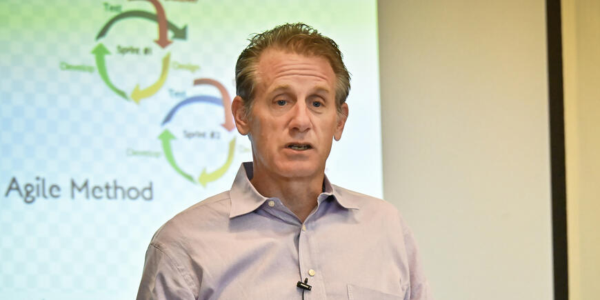 A man in a button-down shirt gives a presentation in front of a projector screen