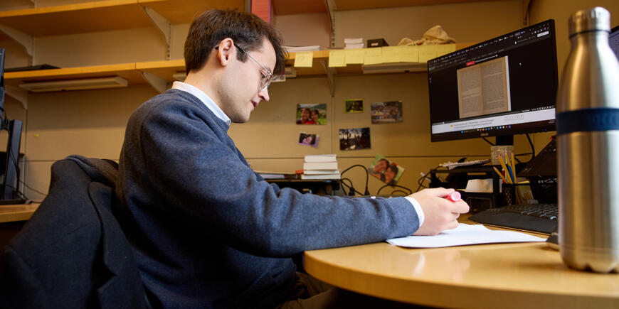 PhD student Alex Busch sits at his desk in his office. There are bookshelves behind him and he is highlighting a stack of papers 