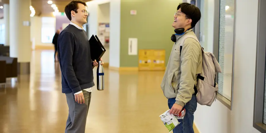 PhD student Alex Busch stands on the left side of the frame facing Soohyun Roh another Phd student. They are talking and smiling