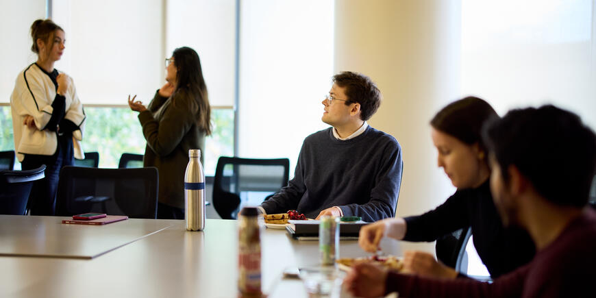 PhD student Alex Busch sits at a large conference room table. Several other people are in the room with him. He is looking up at a presentation off screen