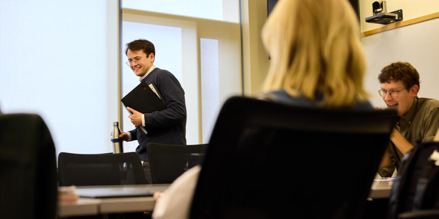 PhD student Alex Busch takes a seat at a table in classroom. Nathan Wilmers sits at the head of the table