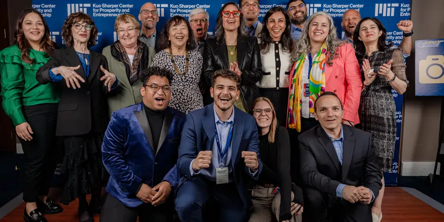 A group of 15 people in business wear, smiling in front of a blue and white MIT backdrop