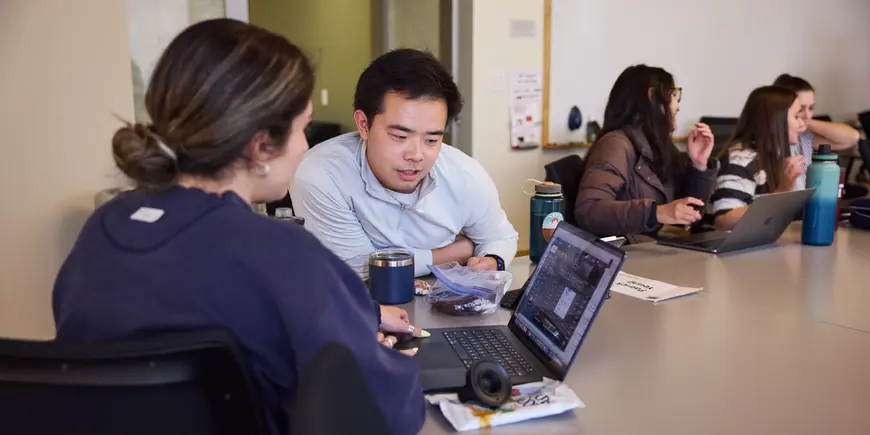 Patrick Yeung sits at a conference table with other students. He and another student look at a laptop together, discussing what they see there