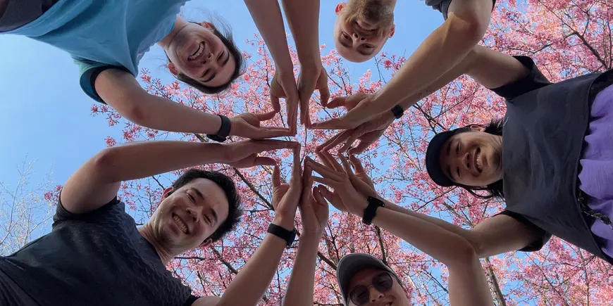 Five members of the MIT Sloan Run club, including Patrick Yeung, bottom left, make a star symbol with their hands. the camera is on the ground and they are looking down at it smiling