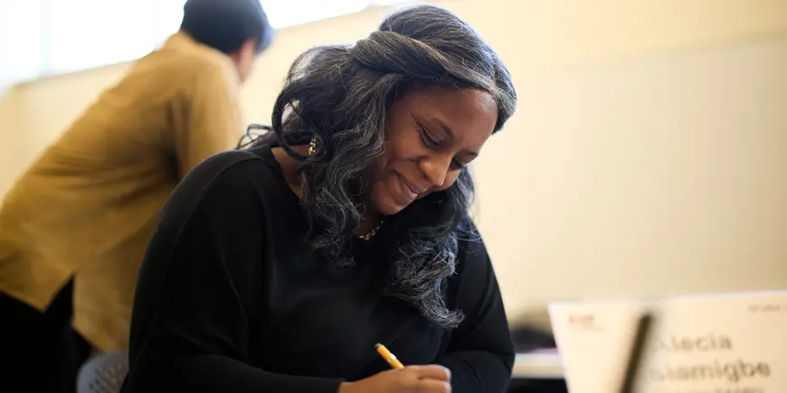 Alecia Asiamigbe smiles down at a paper and pen as she writes something in an MIT Sloan classroom.