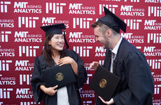 Man and woman in graduation cap and gown each holding their diplomas, facing each other and smiling. A red background with the MIT Master of Business Analytics in white lettering.  