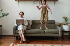 Woman sitting on a sofa and working at a computer while an energetic child jumps on the sofa besider her