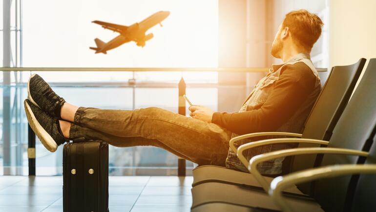 Man sitting in airport waiting area, staring out window at a plane taking off