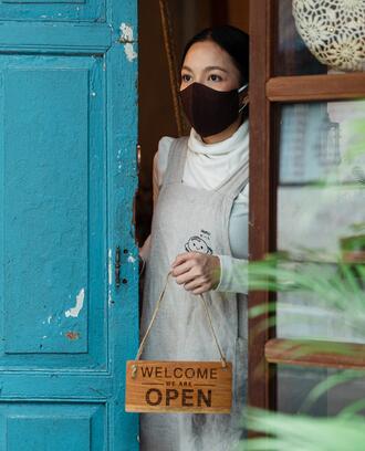 Woman in a face mask opening the door of a business holding a "Welcome, we're open" sign looking tentative and unsure.