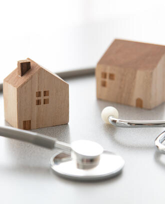 Two mini wooden house figurines with a regular sized stethoscope wrapped around them on smooth white surface