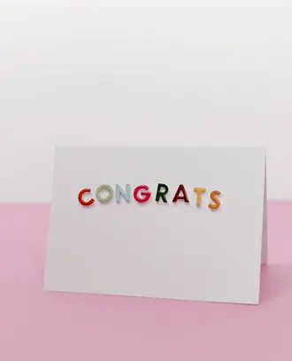 A white card with the word "congrats" on it in colorful letters, against a pink-and-white background