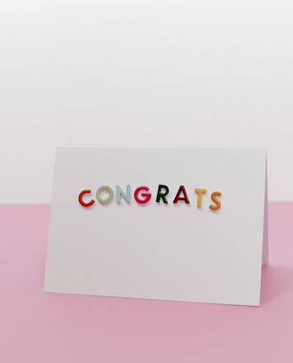 A white card with the word "congrats" on it in colorful letters, against a pink-and-white background