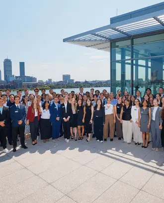 The MBAn Class of 2025 lines up for a photo outside with the Charles River and the Boston skyline in the background