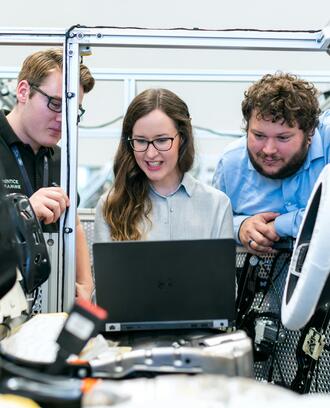 Three people look at a laptop in a some type of industrial setting