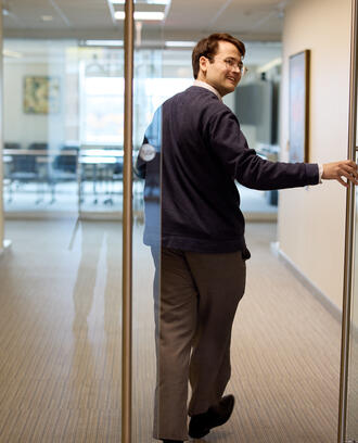 PhD student Alex Busch holds open a door and smiles over his shoulder as he walks through a building at MIT Sloan