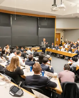 Wide shot of a classroom filled with students facing professor lecturing up front. Behind the professor are empty blackboards.