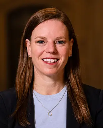 Photo of Megan Greene, a fair-skinned woman with long brown hair and light eyes wearing a dark blazer, a light blue sweater, and a simple necklace, smiling.