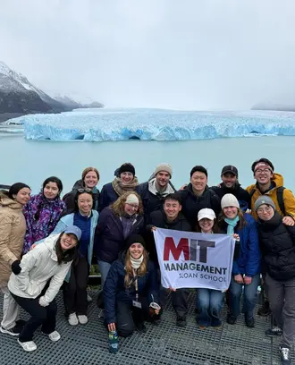 Study tour attendees pose in a group photo in front of a glacier holding an MIT sign