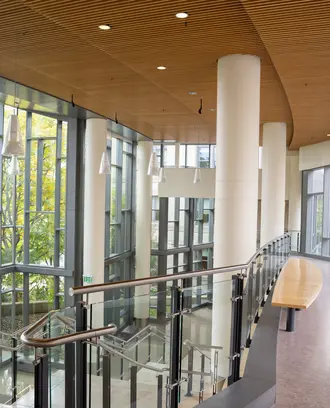 Empty second floor hallway in e62. To the left is a staircase down to the first floor. To the right is the bridge that leads to building E52 if you keep walking straight. There is a woman out of focus walking on the bridge towards the camera all the way to the right of the bridge.