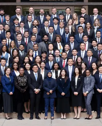 MIT SFMBA Class of 2026 standing on the steps in front of the Memorial Drive Entrance of Building E52 wearing professional business attire.
