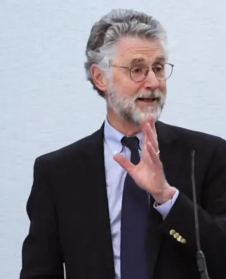 A man with gray hair wearing a dark suit and glasses stands at a podium speaking and gesturing with his hand
