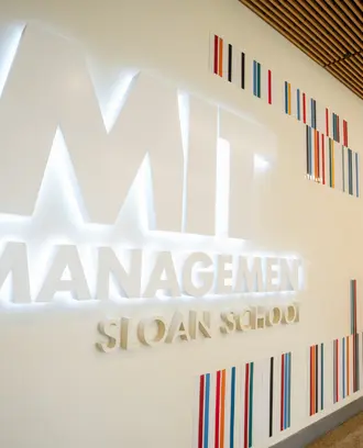 Illuminated MIT Sloan School of Management sign in a campus hallway, with a blurred person walking past.