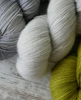 Photo of 3 skeins of yarn: 1 silvery gray, 1 white, and 1 leaf green, sitting on a gray wooden surface