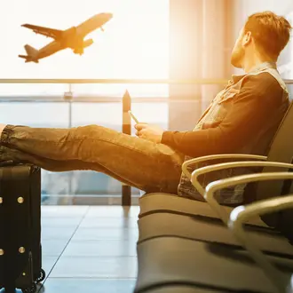 Man sitting in airport waiting area, staring out window at a plane taking off