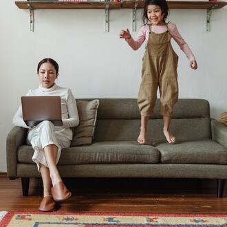 Woman sitting on a sofa and working at a computer while an energetic child jumps on the sofa besider her